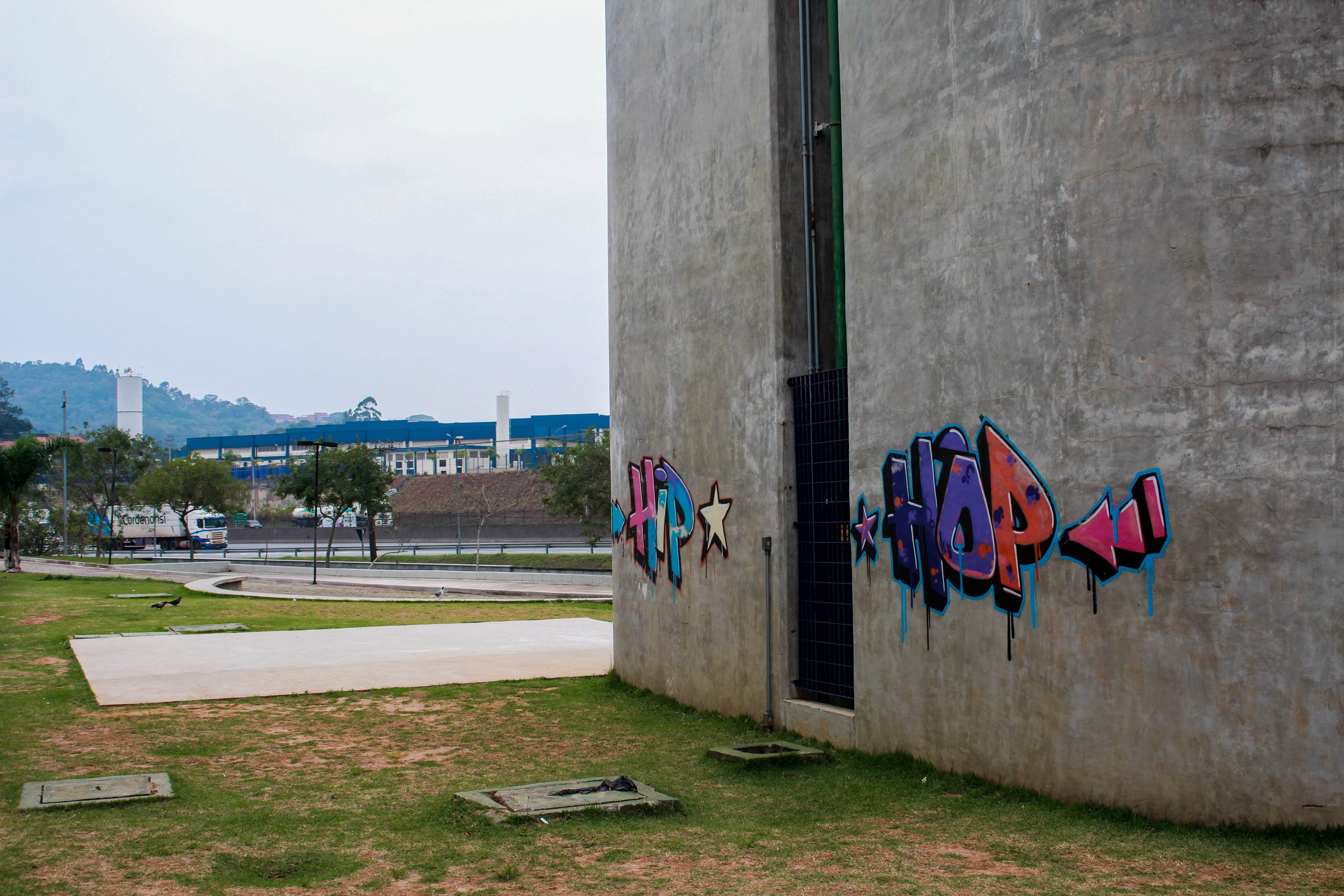 Betongebäude mit Graffiti an seiner Seite, umgeben von Gras, Bäumen, Strommasten, Fahrzeugen auf der Straße, anderen Gebäuden, Hügeln und einem klaren blauen Himmel.