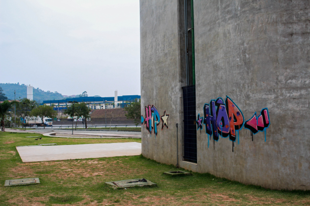 Betongebäude mit Graffiti an seiner Seite, umgeben von Gras, Bäumen, Strommasten, Fahrzeugen auf der Straße, anderen Gebäuden, Hügeln und einem klaren blauen Himmel.