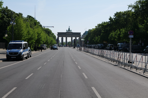 Ein Polizeifahrzeug steht auf der Seite einer vielbefahrenen Straße vor dem Brandenburgertor in Berlin, Deutschland, mit Barrieren, Schildern, Bäumen und Laternen im Hintergrund und einer bewölkten Himmel.