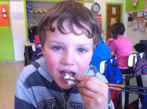 Ein Junge in einer grauen und schwarzen Jacke isst mit Stäbchen in einem Klassenraum, hinter ihm sitzen andere Kinder, grüne und weiße Wände, Lehrplakate, eine braune Tür und eine Wanduhr sind zu sehen.