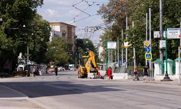 Eine Stadtstraße mit einer Baustelle in der Mitte, mit Fahrzeugen, Fußgängern, einem Radfahrer, Verkehrsleitkegeln, Pfosten, Hinweisschildern, Strommasten mit Drähten, Bäumen, Gebäuden mit Fenstern und einem bewölkten Himmel im Hintergrund.