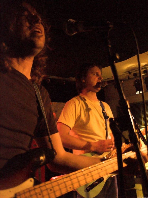 Ein Musikkonzert mit zwei Sängern: einer in einem weißen Hemd, der eine grüne Gitarre hält, und ein anderer in einem schwarzen Hemd mit Brille, der eine schwarze Gitarre hält, mit Scheinwerfern und anderen Instrumenten im Hintergrund.