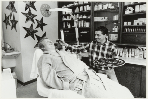 Ein Mann lächelt in einem Zahnarztstuhl, während ein anderer Mann neben ihm steht und ein Tablett hält, mit einem Waschbecken, Sternen, einer Uhr und Regalen voller Flaschen im Hintergrund.