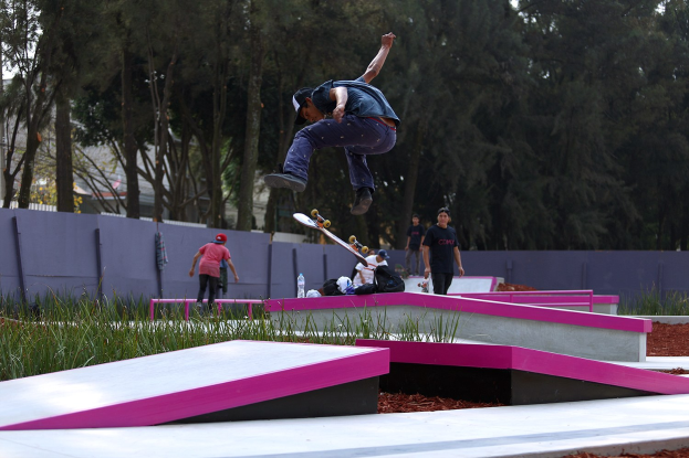 Ein Skateboarder führt einen Sprung auf einer Rampe aus, während Zuschauer in der Nähe stehen, einige halten Skateboards, mit verstreuten Gegenständen und einem Hintergrund aus Bäumen, Gebäuden und einem klaren Himmel.