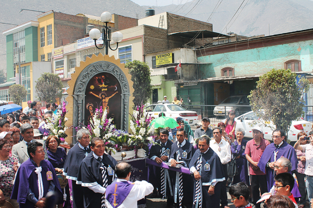 Menschen auf einer Straße vor der Stadt, mit einem Kreuz und Blumensträußen im Vordergrund, fahrende Autos im Hintergrund und Gebäude, Bäume und Straßenlaternen weiter hinten.