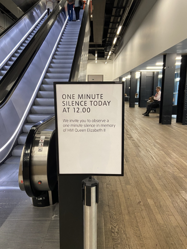 Eine Rolltreppe im Flughafen mit einem Schild, auf dem "Eine Minute Stille heute" steht, sowie ein paar Menschen darauf und Lampen an der Decke im Hintergrund.
