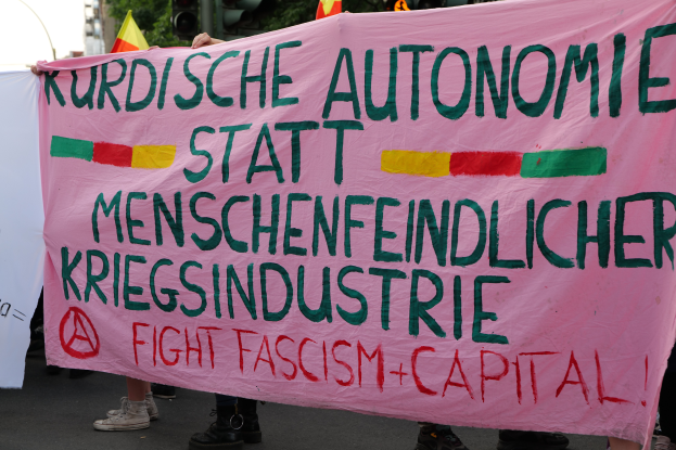 Eine Gruppe von Menschen steht auf einer Straße und hält ein pinkes Banner hoch, auf dem "Kurdische Autonomie Statt Menschenfeindlicher Kriegsindustrie Fight Fascism Capital" steht, mit Verkehrszeichen, Flaggen, Bäumen, Gebäuden und einem klaren blauen Himmel im Hintergrund.