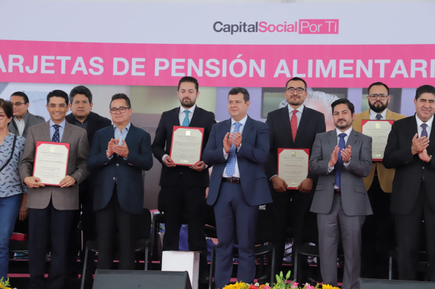 Gruppe von Menschen auf einer Bühne klatschend mit Zertifikaten in den Händen, mit Stühlen in einem Halbkreis angeordnet, einem Banner mit der Aufschrift "Capital Social for Ti" im Hintergrund und Blumensträußen und Lautsprechern unten.