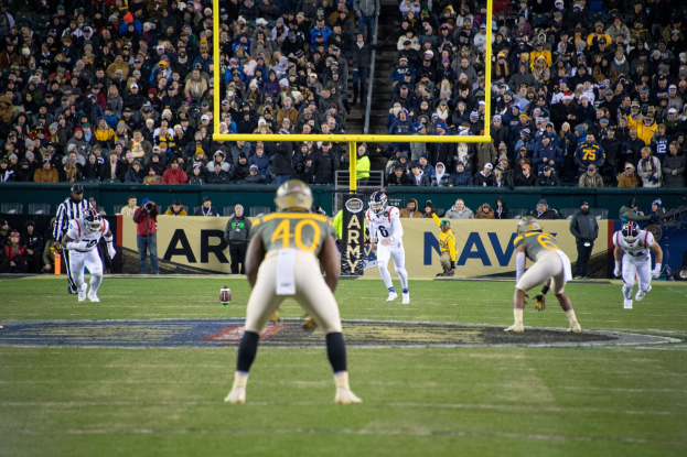 Ein Fußballspiel im Gange mit Spielern in Helmen und Sportbekleidung im Vordergrund und einer Zuschauermenge im Hintergrund, mit sichtbaren Pfosten und Tafeln mit Text.