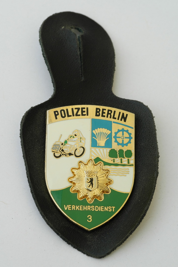 Ein schildförmiges Polizeibadgebnis mit einem blauen Hintergrund, weißer Umrandung und einem zentralen weißen Stern, beschriftet mit 'Polizei Berlin' in schwarzer fettgedruckter Schrift, das auf einer Oberfläche ruht.