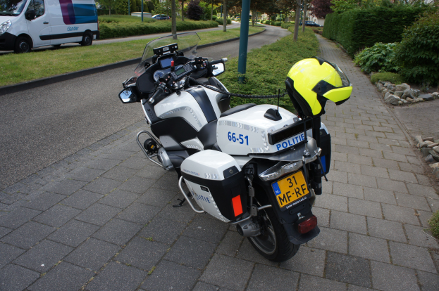Ein auf der Seite der Straße geparktes Polizeimotorrad mit einem Helm darauf, umgeben von Bäumen, Masten, Gebäuden, Pflanzen, Gras, Steinen und anderen Fahrzeugen.