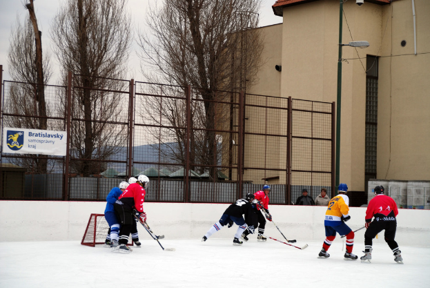 Menschen beim Eisschnellaufen auf einer Eisbahn mit Gebäuden, Bäumen, einer Straßenlaterne, einem Namensschild und Zäunen im Hintergrund unter einem Himmel.