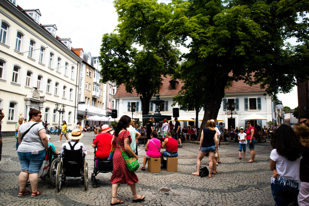 Eine Gruppe von Menschen, einige im Rollstuhl, geht eine Kopfsteinpflasterstraße in der Altstadt von Heidelberg entlang, mit Bäumen, Gebäuden, Laternen und einer Statue im Hintergrund unter einem bewölkten Himmel.