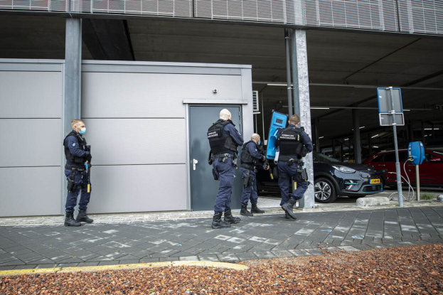 Eine Gruppe von Polizisten steht in einer Parkplatz vor einem Gebäude mit einer Tür, umgeben von Fahrzeugen, Pfählen, Plakaten und verstreuten Steinen.