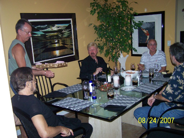 Eine Gruppe von Menschen an einem Esstisch mit Weingl√§sern, Flaschen und einer Sch√∂pfel mit Essen, ein Mann bedient, Fotos auf dem Wand und eine Pflanze auf der Seite.