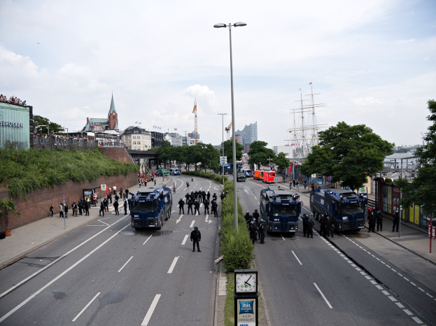 Eine große Gruppe von Polizeibeamten steht an der Seite einer Straße, umgeben von Fahrzeugen und Menschen, mit Laternen, Schildern mit Text, Pflanzen, Bäumen, Gebäuden und Türmen im Hintergrund unter einem bewölkten Himmel.
