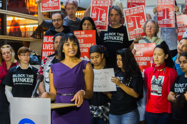 Eine Black Lives Matter-Aktivistin spricht an einem Podium mit Mikrofon zu einer Menge mit Schildern, während im Hintergrund Lichter und Gegenstände zu sehen sind.