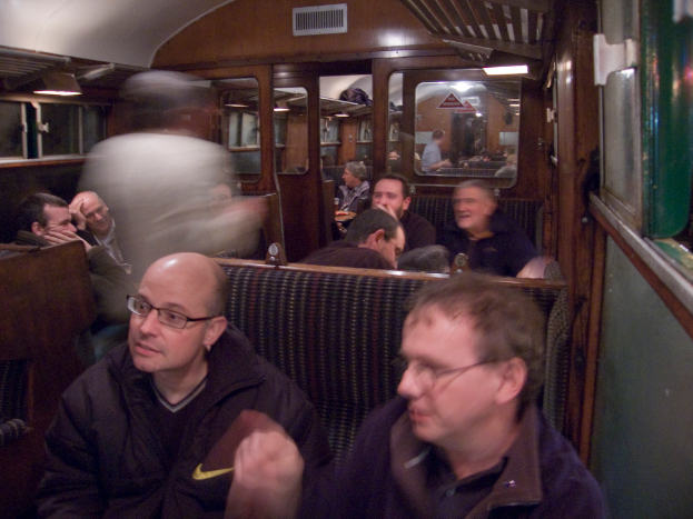 Zwei Männer sitzen auf einem Sitz in einem Waggonabteil, hinter ihnen ist eine Gruppe von Menschen zu sehen; ein Eingang zwischen den Abteilen ist oben zu sehen und ein Fenster rechts.