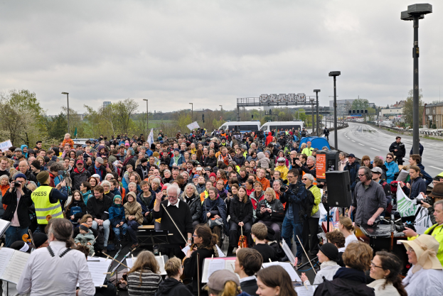 Eine große Gruppe von Menschen marschiert vor einer Menge, einige halten Musikinstrumente und Bücher mit Ständern, mit Fahrzeugen, Laternenpfählen, Bäumen, Gebäuden und einem bewölkten Himmel im Hintergrund.