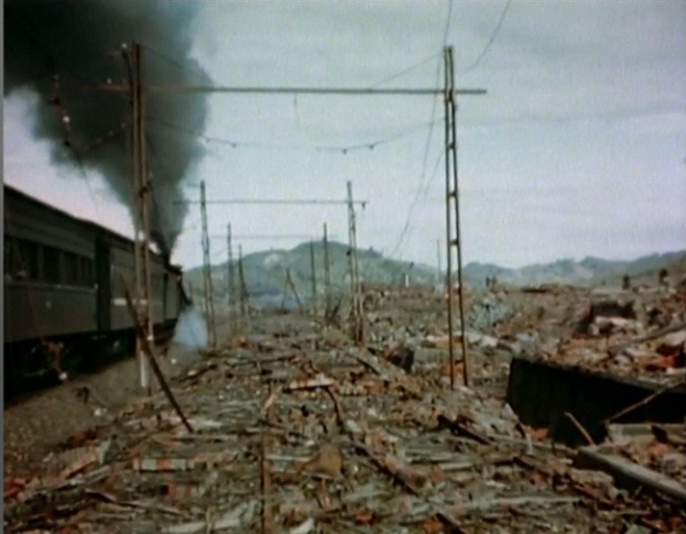 Ein Zug fährt durch ein beschädigt Gebiet mit Rauch, der aus ihm aufsteigt, umgeben von Masten und Drähten, Höhen und einem klaren Himmel im Hintergrund und verstreuten Trömmern am Boden.