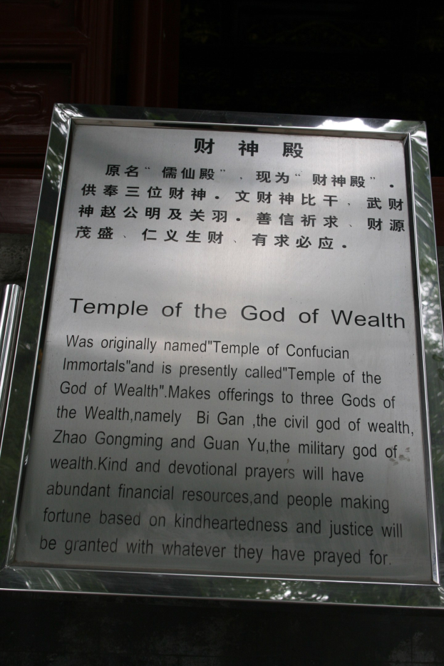 Ein Tempel, der dem Gott des Reichtums gewidmet ist, mit einer Namensplakette im Vordergrund und einer Tür im Hintergrund, umgeben von üppiger Vegetation.