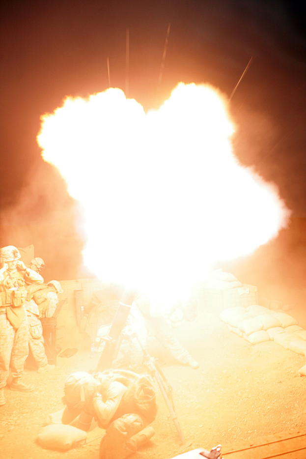 Mehrere Personen in Uniformen und Helmen befinden sich auf einem Geländebereich mit einigen Taschen drumherum, wobei in der Mitte des Bildes ein Feuer brennt.