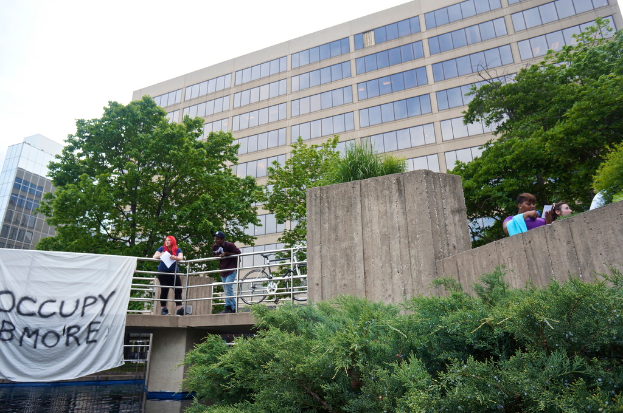 Menschen auf einer Brücke mit einem Banner und einem Fahrrad, unter dem Wasser fließt, mit Bäumen, Gebäuden und Himmel im Hintergrund.