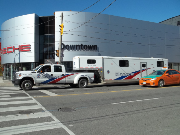 Ein Polizei-Lkw vor einem Gebäude mit Text, eine Ampel mit Mast und Drähten im Vordergrund und Bäume mit einem klaren blauen Himmel im Hintergrund.