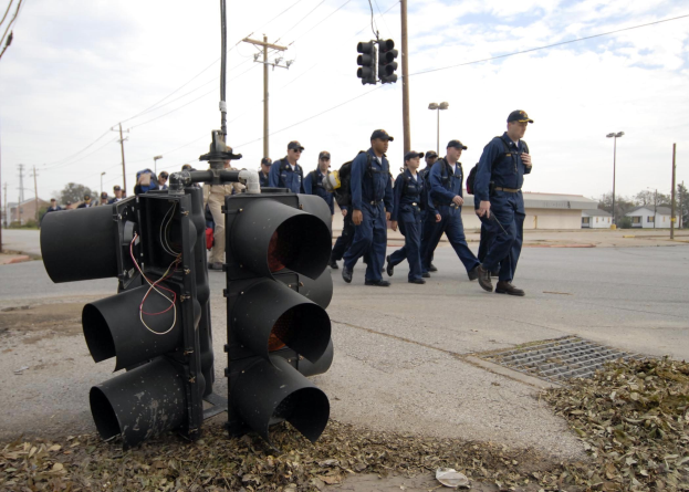 Polizisten in Uniform und Mützen auf einer Straße mit Strommasten, Ampel, Gebäuden und Bäumen unter einem klaren blauen Himmel.