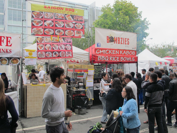 Menschen stehen auf einer Straße außerhalb der Stadt, ein Mann in einem grauen und weißen T-Shirt hält eine Wasserflasche und eine Frau in einer blauen Jacke einen Kinderwagen. Im Hintergrund sind Stände mit weißen und roten Zelten, farbige Banner und Gebäude, Bäume und eine Straßenlaterne zu sehen.