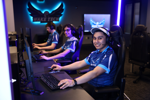 Eine Gruppe von Menschen sitzt an Schreibtischen mit Computern, trägt Headsets und lächelt, in einem Raum mit Monitoren, Tastaturen und einem Logo an der Wand.