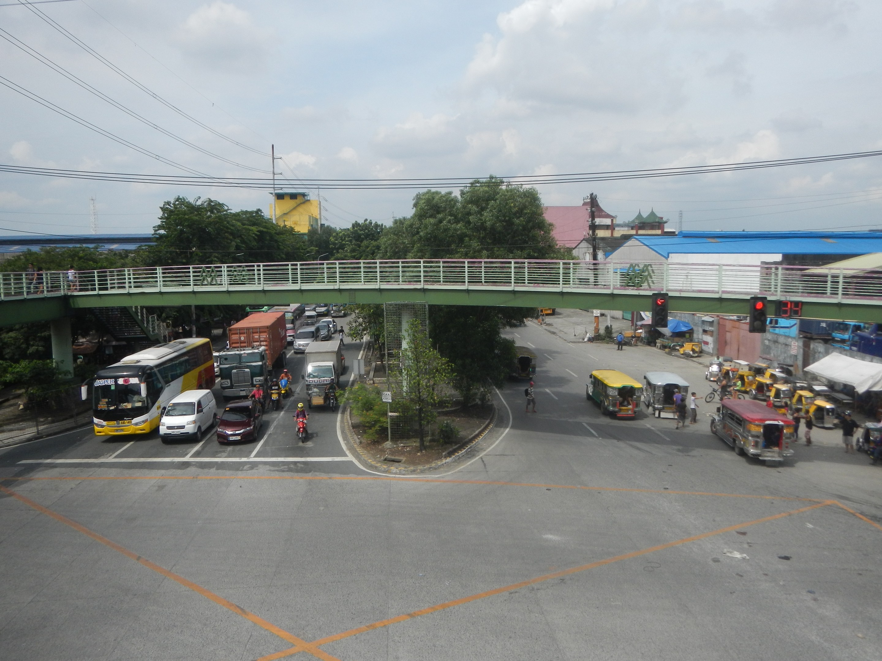 Eine belebte Stadtstraße mit Autos, Bussen, Fußgängern auf einem Gehweg, einer Brücke mit Geländern, Verkehrsampeln, Bäumen, Gebäuden, Strommasten mit Drähten und einem bewölkten Himmel.