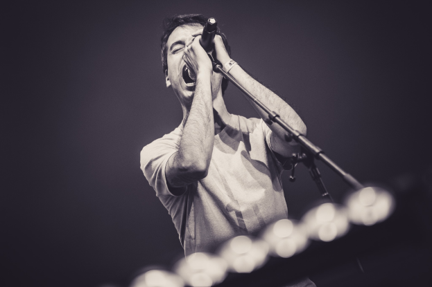 Ein Mann singt in ein Mikrofon auf einer hell erleuchteten Bühne, trägt ein schwarzes T-Shirt und Jeans und hat seine Augen geschlossen und seine Haare wild gestylt.