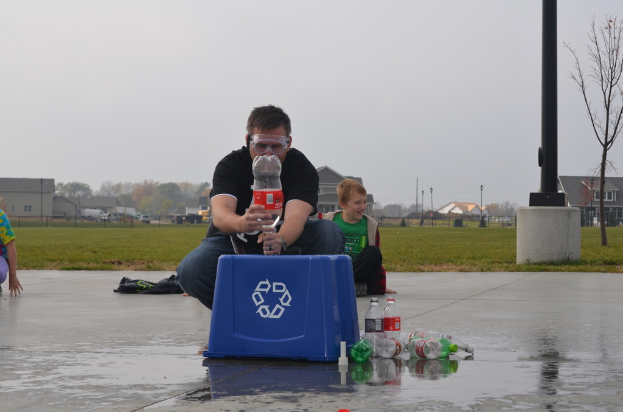 Ein Mann mit Brille hält eine Flasche und steht zwischen mehreren Flaschen und einer Kiste, umgeben von Pfählen, Bäumen, Gebäuden, Fahrzeugen und Gras.