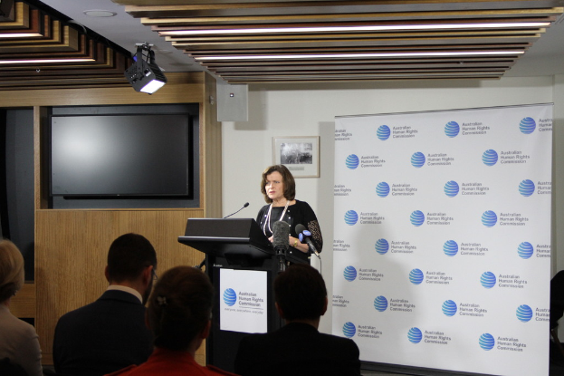 Eine Frau steht auf einer Bühne und spricht in ein Mikrofon, vor ihr sitzen Menschen auf Stühlen, hinter ihr hängt ein Banner an der Wand, ein Fernseher an der Holzwand und Lampen an der Decke.