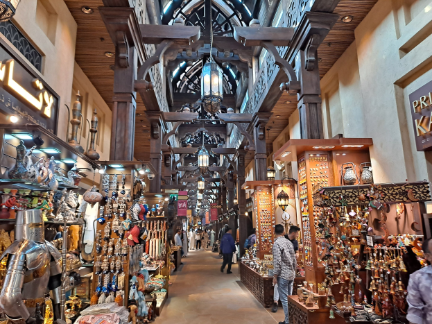 Ein geschäftiges Dubai-Basar mit Menschen, die zwischen Regalen voller Gegenstände hindurchgehen, Wänden mit Text und einer Decke mit warmem Licht.