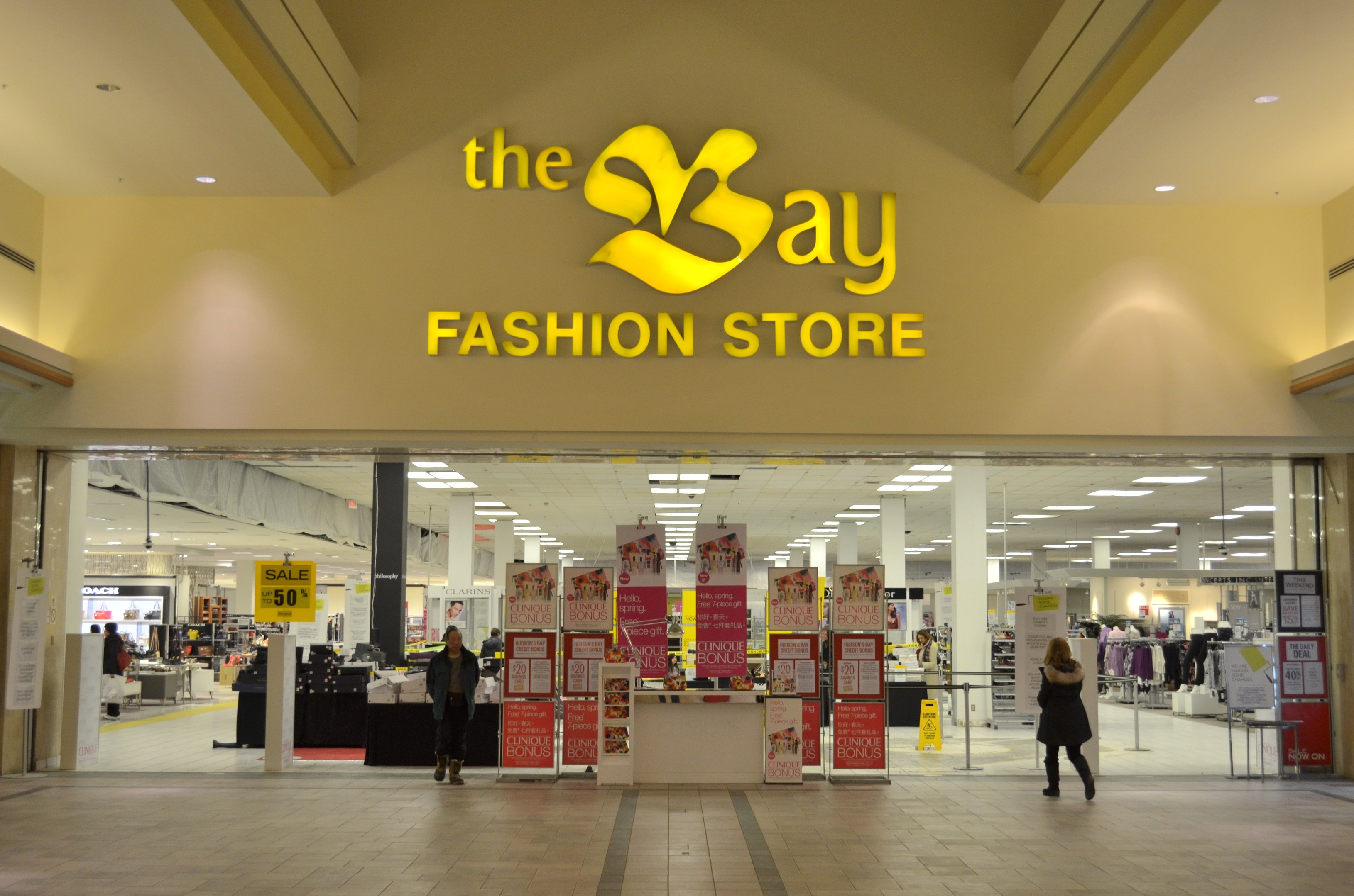 Außenansicht eines Bay Fashion Stores in einem Einkaufszentrum mit Einkäufern, Wandtafeln, Säulen, Deckenleuchten und Hintergrundauslagen.