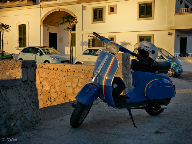 Ein Haus mit drei parkenden Autos davor, eine kleine Wand neben den Autos mit einem Roller daneben, bei hellerer Sonne.