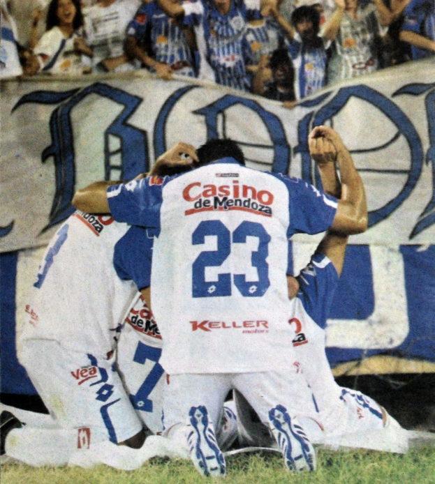 Gruppe von Fußballspielern, die auf dem Boden liegen und mit jubelnden Zuschauern feiern, im Hintergrund ist eine Fahne mit Text zu sehen.