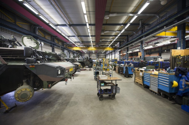 Ein großer Lagerraum voller Maschinen, darunter Fahrzeuge auf der linken Seite, Wagen mit Gegenständen, Holzboxen und andere Gegenstände auf der rechten Seite, mit Lichtern oben.