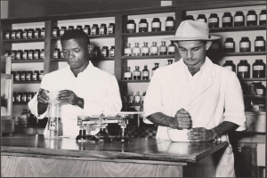 Zwei Männer in weißen Kitteln und Hüten stehen an einem Tresen in einer Apotheke mit einer Waage, einer Schüssel und Regalen mit Flaschen im Hintergrund, dargestellt in Schwarz-Weiß.