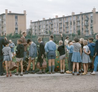 Kinder, die vor einem Gebäude mit blühenden Pflanzen, Bäumen und einem klaren blauen Himmel stehen, einige mit Schulranzen.