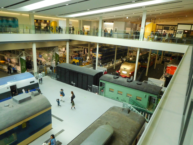 Ein großer Museumsraum mit verschiedenen Arten von Zügen, mit Menschen, die beobachten, Geländern, Säulen, Deckenleuchten und Informationsschildern.