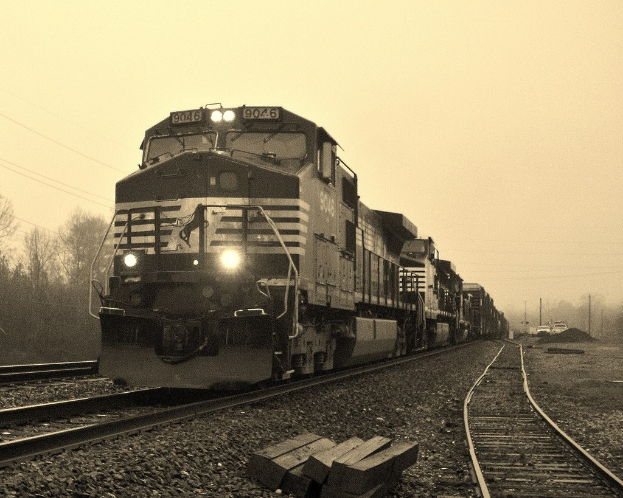 Ein Zug auf Schienen mit Autos, Strommasten, Bäumen und Himmel.
