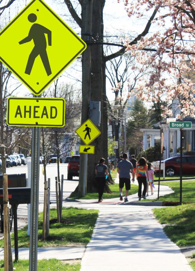 Eine Gruppe von Menschen, die auf einem Bürgersteig neben einem gelben Fußgängersignal gehen, mit Gras, Bäumen, Fahrzeugen, Gebäuden und einem klaren blauen Himmel im Hintergrund.