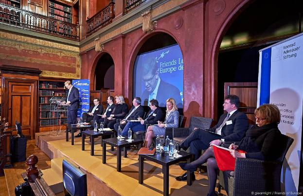 Ein Mann steht an einem Podium und spricht zu einem Publikum, das auf einer Bühne sitzt. Im Hintergrund sind Wasserflaschen auf einem Tisch, ein Projektionsschirm, ein Banner und ein Bücherregal zu sehen.