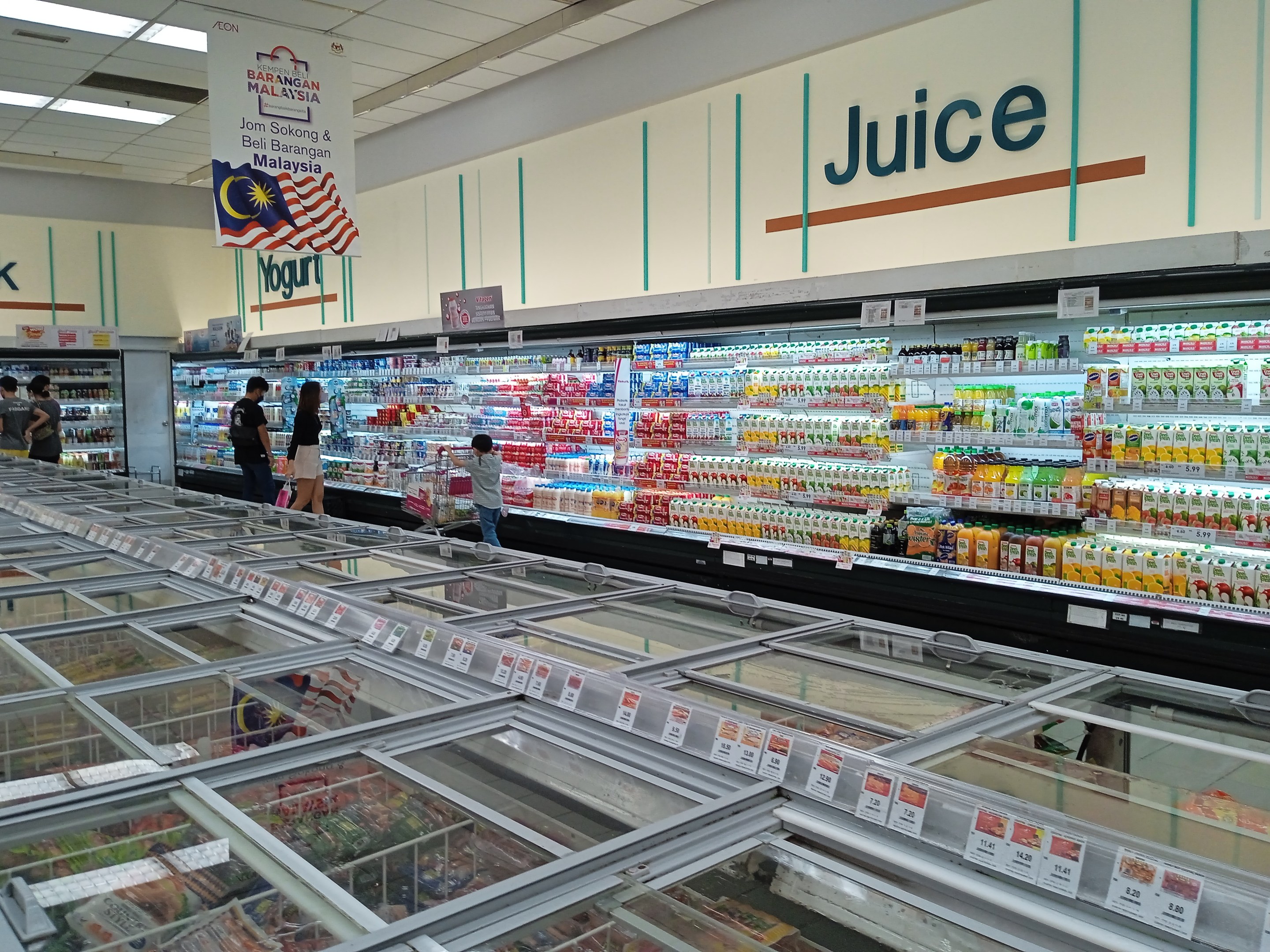 Ein hell erleuchteter Saftladen in einem Supermarkt zeigt eine Reihe von Flaschengetränken, frischem Gemüse und anderen Lebensmitteln auf Regalen, mit ein paar Kunden, die stöbern und Deckenleuchten, die den Raum erhellen.