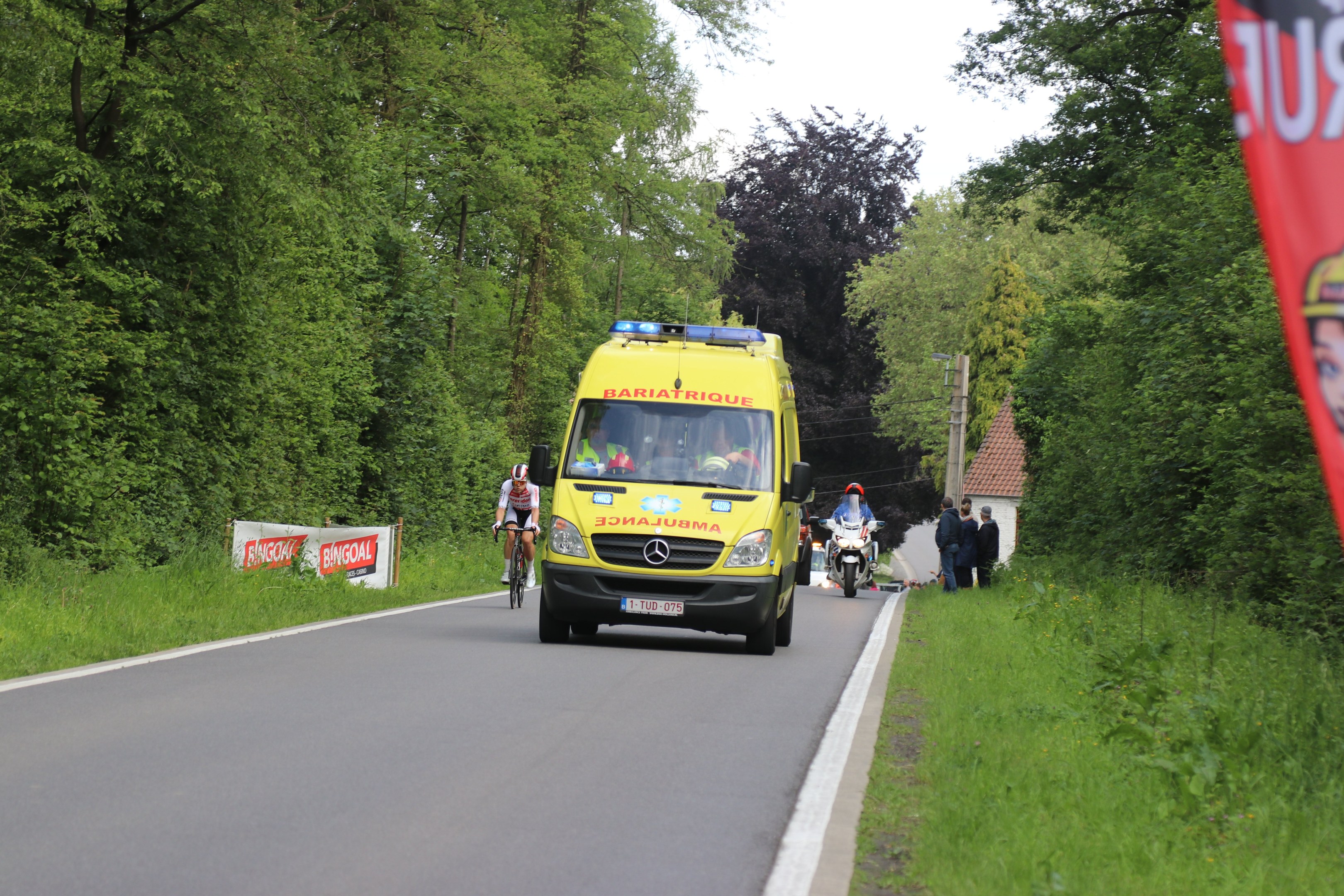 Ambulanz fährt auf einer Straße mit Radfahrern äquidistant, Gras und Bäume auf beiden Seiten, Häuser, Masten und einen klaren blauen Himmel im Hintergrund.