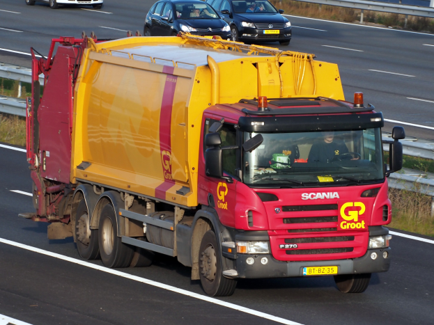 Ein gelb-roter Müllwagen fährt auf einer Autobahn, umgeben von anderen Fahrzeugen, mit Gras und einer Begränzung auf beiden Seiten der Straße.