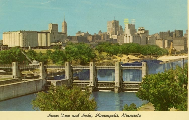 Eine Brücke überspannt einen Fluss mit Bäumen und Gebäuden im Hintergrund, mit dem Text "Lower Dam and Locks, Minneapolis, Minnesota" unten.